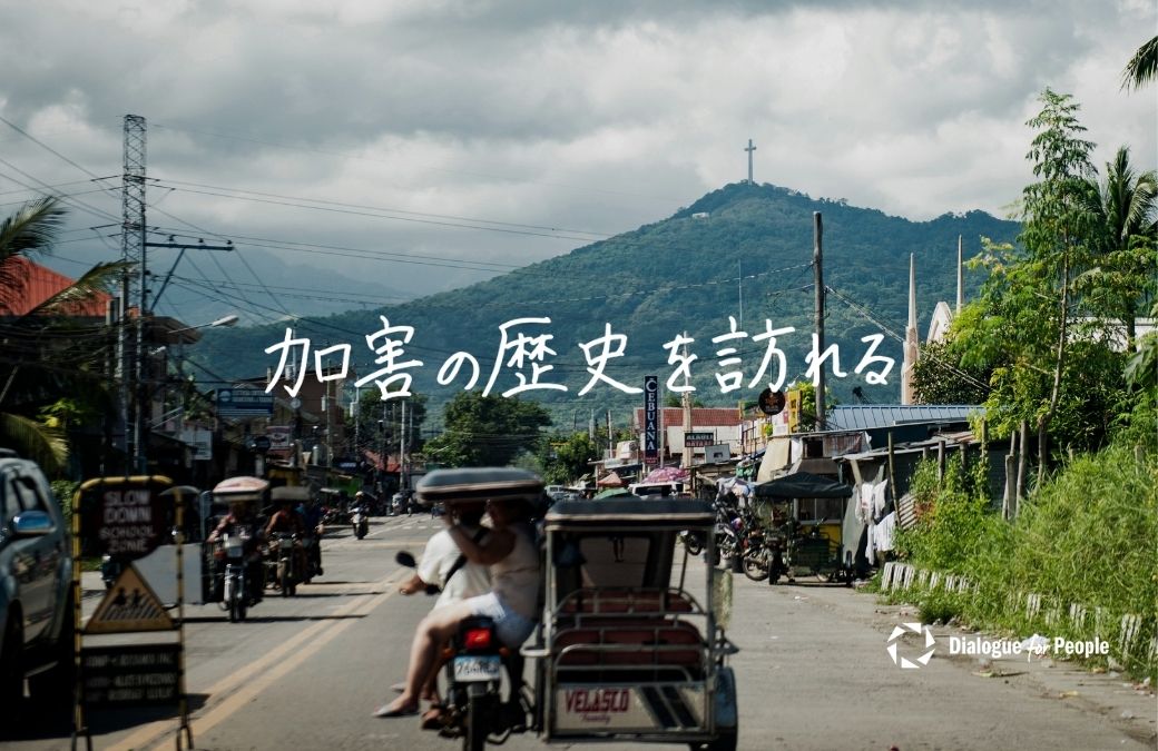 加害の歴史を訪れる。フィリピンの街並み、走る乗り物、そして奥の山頂に立つ十字架の風景