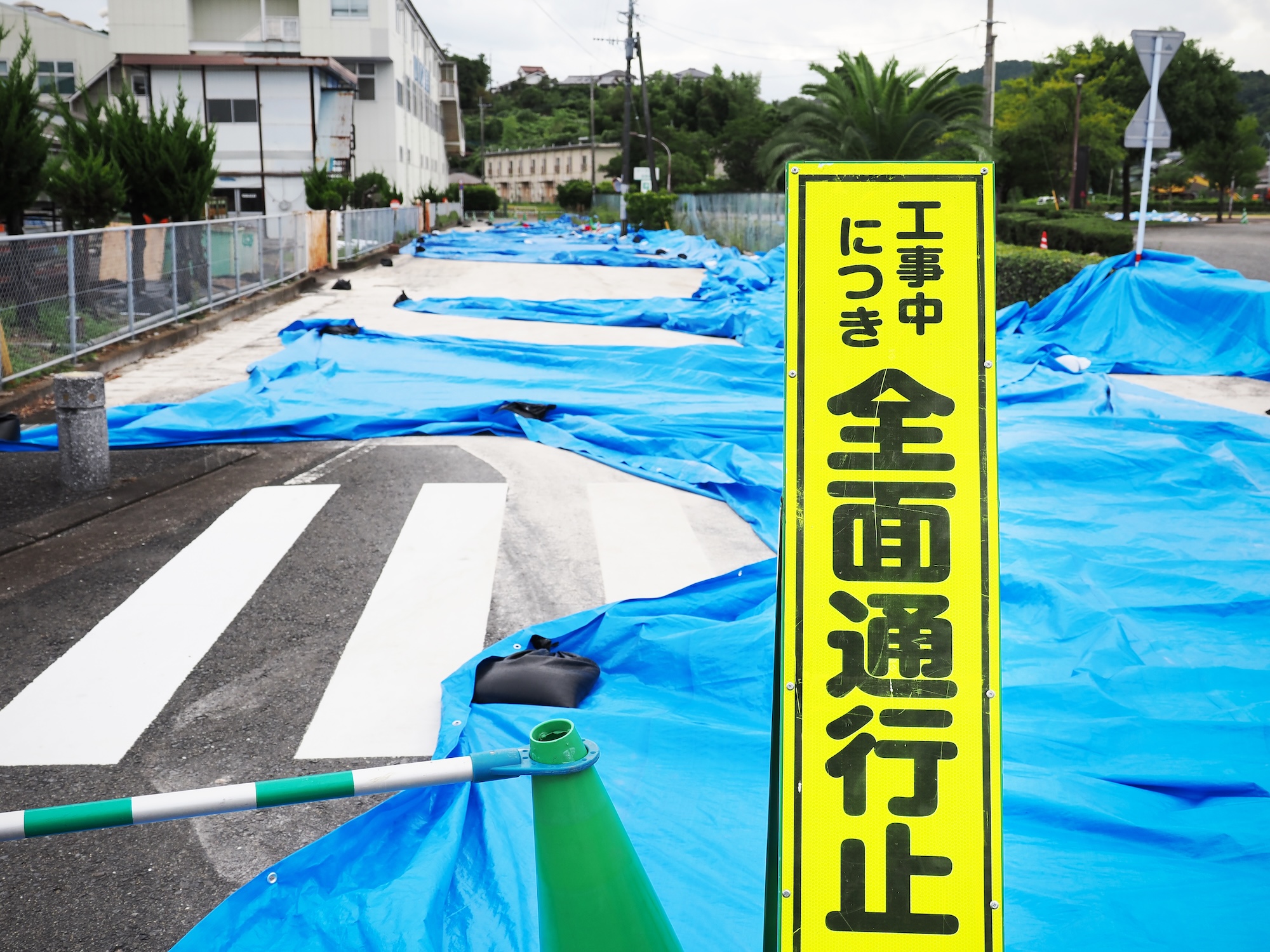 手前に「工事中につき 全面通行止」と書かれた黄色の看板が立ち、奥の道路全体に青いシートが敷き詰められた市道の様子