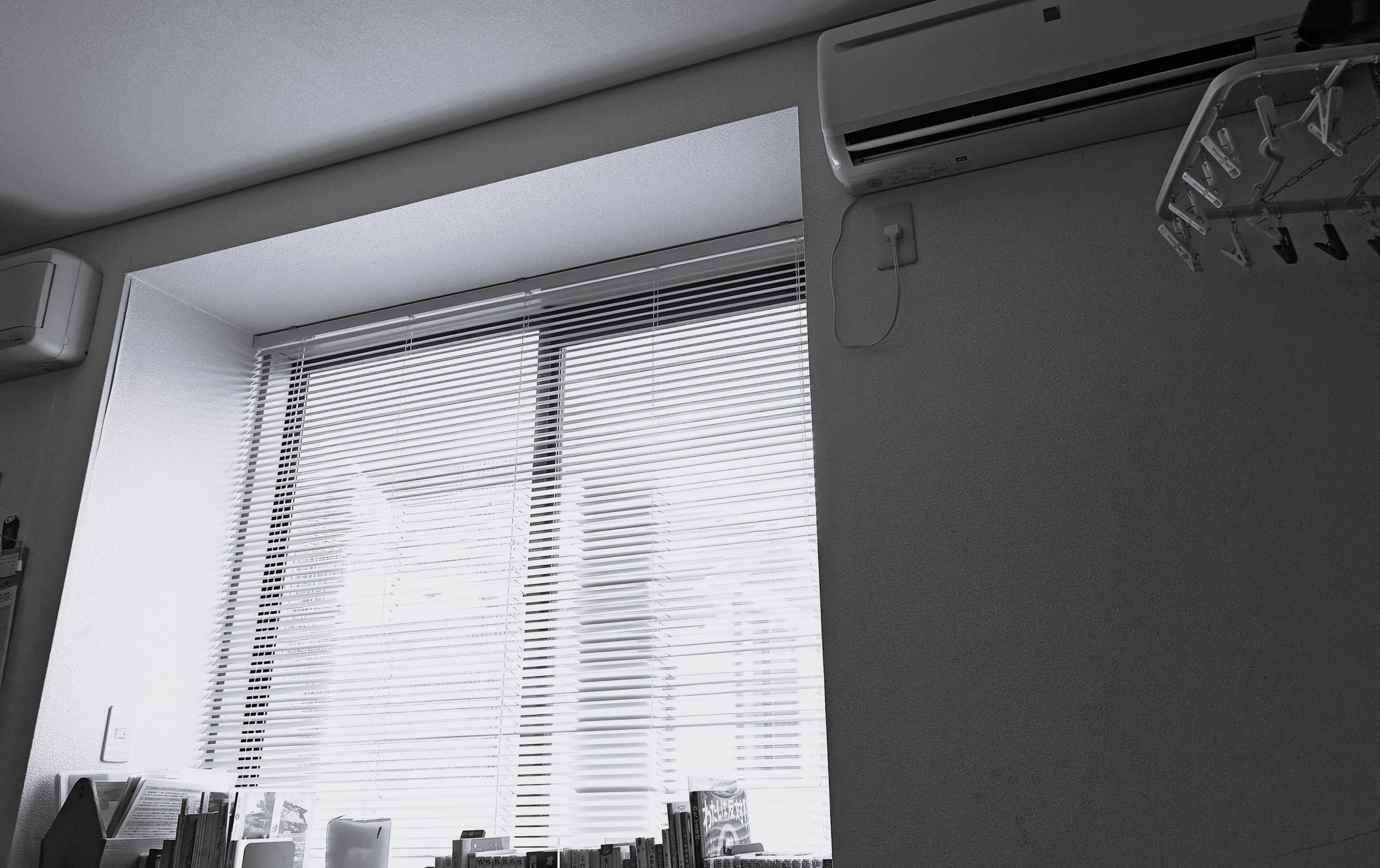 モノクロの写真。ブラインドが下りた大きな窓のある室内。壁にはエアコンやハンガーが掛けられている。