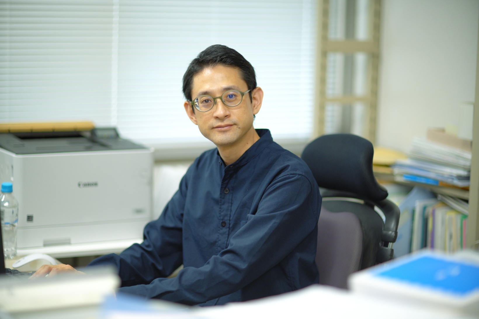 宮城教育大学の研究室にて、デスクの前に座りカメラに向かって微笑む松﨑丈さん。メガネをかけ、紺色の長袖シャツを着用。背景にはプリンターや本棚が並んでいる。
