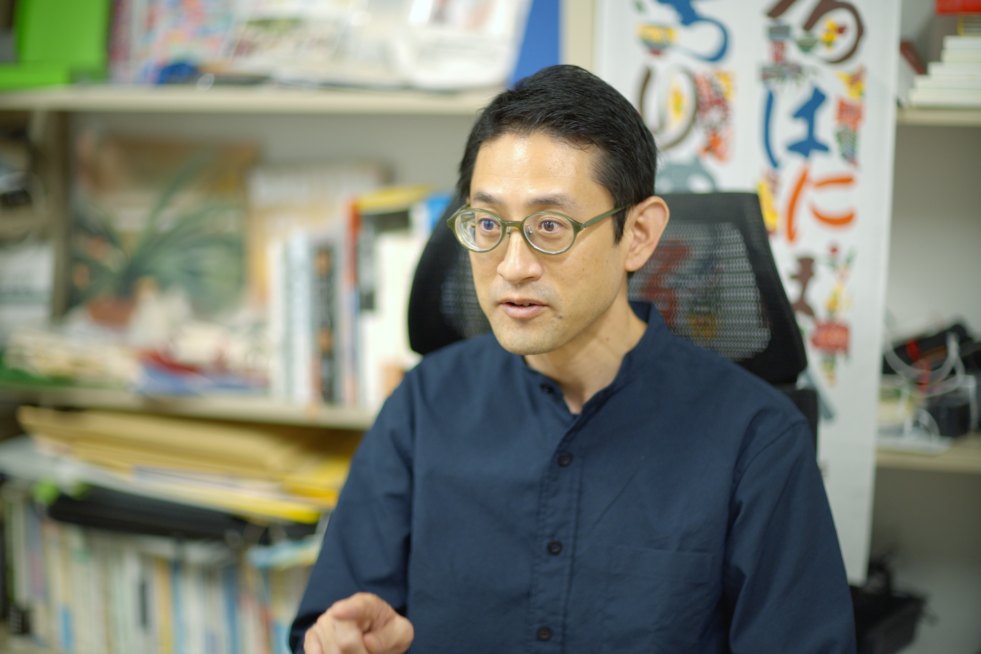 インタビューに応じる松﨑丈さん。メガネをかけ、紺色の服を着て、手話またはジェスチャーを交えながら語っている姿。