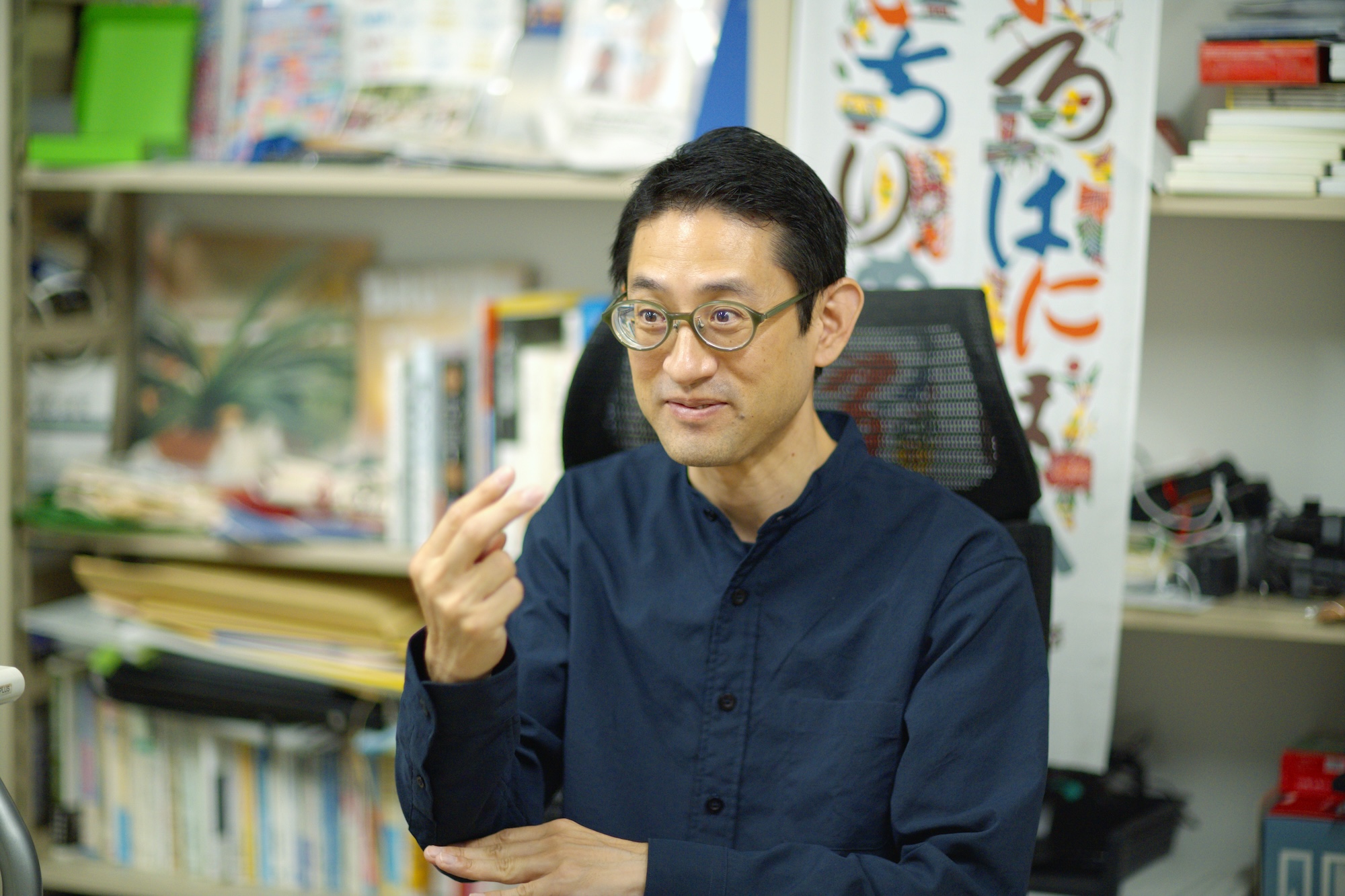 手話を交えて質問に答える松﨑丈さん。メガネをかけ、紺色の長袖服を着て話している様子。